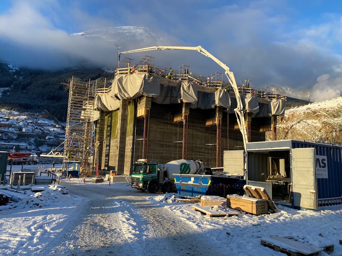 Byggeplassen med snø på bakken og en betongbil som leverer betong til en pumpe.